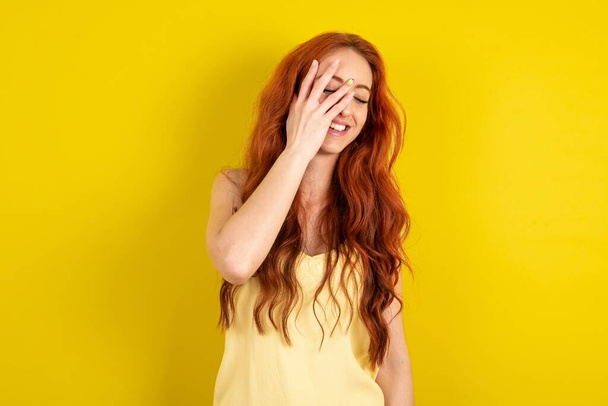 young red-haired wearing yellow t-shirt yellow studio background makes face palm and smiles broadly, giggles positively hears funny joke poses - Photo, image