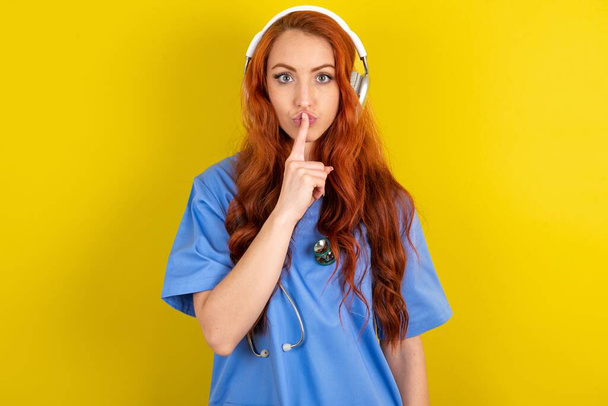 giovane donna dai capelli rossi medico su sfondo giallo studio facendo gesto di silenzio con le dita sulle labbra indossando cuffie wireless. Sta 'zitto.. - Foto, immagini
