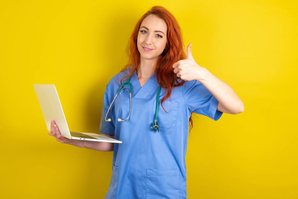 piękne atrakcyjne młody rudowłosy lekarz kobieta nad żółty studio tło trzymać netbook strony palec pokazać kciuk w górę - Zdjęcie, obraz