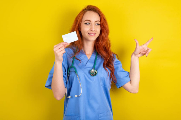 Curioso sorrindo jovem ruiva médico mulher sobre amarelo estúdio fundo mostrando plástico banco mostrando dedo copyspace - Foto, Imagem
