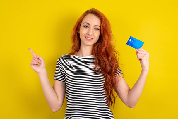Smiling young red-haired wearing striped t-shirt yellow studio background showing debit card pointing finger empty space - Photo, Image