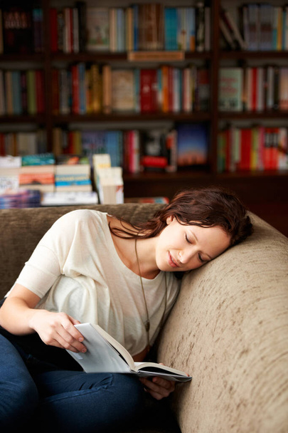 Calm, library and woman reading a book on a sofa for literature while relaxing with shelves. Rest, happy and female person with a smile enjoying a fantasy story or novel on couch in bookstore or shop. - Фото, изображение