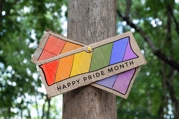 Poteau indicateur qui a dessin arc-en-ciel avec des textes "Mois de la fierté heureuse" collé sur tronc d'arbre dans le parc public pour célébrer et saluer les événements des personnes LGBT dans le mois de la fierté et de respecter la diversité des sexes. - Photo, image
