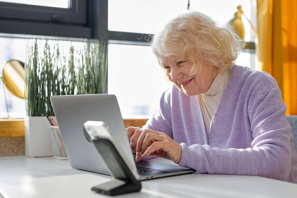 Beautiful senior woman sitting alone, using modern laptop, going to have video call, attending video conference or webinar, typing messages, writing an email. pretty grandmother at home - Фото, зображення