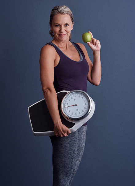Είναι τρόπος ζωής. Studio πορτρέτο μιας ελκυστικής ώριμης γυναίκας που κρατά ένα μήλο και μια ζυγαριά σε μπλε φόντο - Φωτογραφία, εικόνα