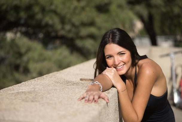Portret van een mooie jonge vrouw met recht bruin haar, leunend tegen een stenen muur kijkend naar de camera glimlachend en gelukkig. Concept mode, schoonheid, trend, geluk, millennial. - Foto, afbeelding