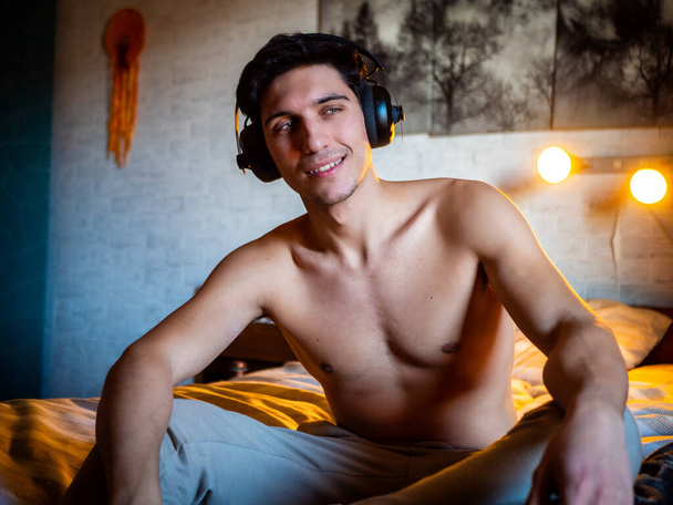 Attractive Young Man Using Headphones Lying Alone On His Bed, Listening to Music and Having a Good Time, Looking at Camera - Zdjęcie, obraz