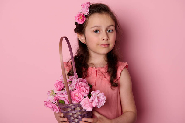 Close-up portrait of beautiful Caucasian blue eyed little girl in stylish pink summer dress, holding a purple basket full of fresh pink roses, isolated background. Happy Childrens Day. Womens Day - Zdjęcie, obraz