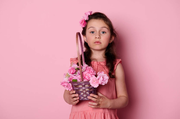 Studio portrait of a Caucasian noble cute little girl with a curly hair and beautiful blue eyes, dressed in a pink dress, holding a basket of roses, looking insightfully at camera over pink background - Фото, изображение