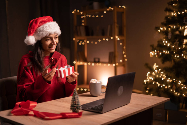 Joven mujer feliz abriendo regalo de Navidad mientras que tiene videollamada sobre un ordenador portátil en la noche en casa. Copiar espacio - Foto, Imagen