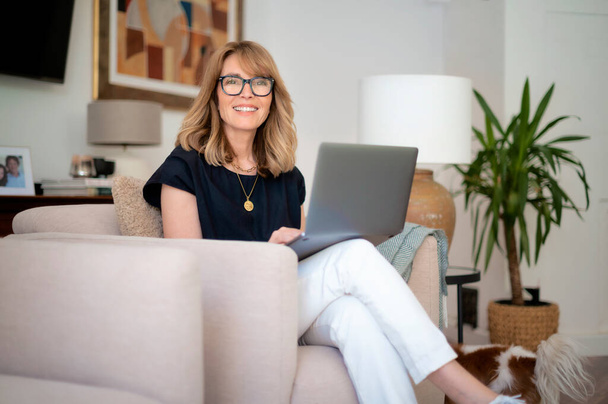 Portrait of attractive middle aged woman sitting at home and using laptop. Blond haired female wearing eyeglasses and black shirt. Home office. - Foto, afbeelding
