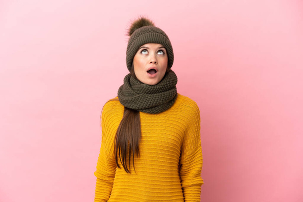 Joven chica caucásica con sombrero de invierno aislado sobre fondo rosa mirando hacia arriba y con expresión sorprendida - Foto, Imagen