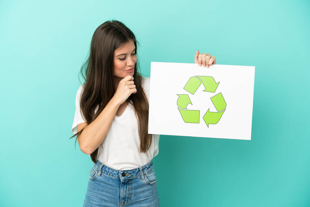 Mladá běloška žena izolované na modrém pozadí drží plakát s ikonou recyklace - Fotografie, Obrázek