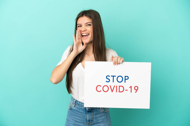 Mulher caucasiana jovem isolado no fundo azul segurando um cartaz com texto Stop Covid 19 e gritando - Foto, Imagem