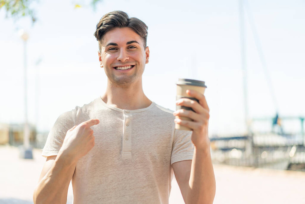 Молодой привлекательный мужчина держит кофе на открытом воздухе с неожиданным выражением лица - Фото, изображение
