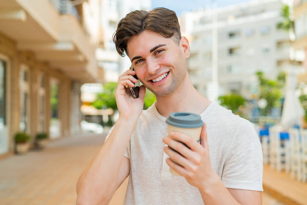 Молодой красивый мужчина, пользующийся мобильным телефоном и держащий кофе со счастливым выражением лица - Фото, изображение