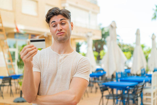 Młody przystojny mężczyzna trzyma kartę kredytową na świeżym powietrzu ze smutnym wyrazem twarzy - Zdjęcie, obraz