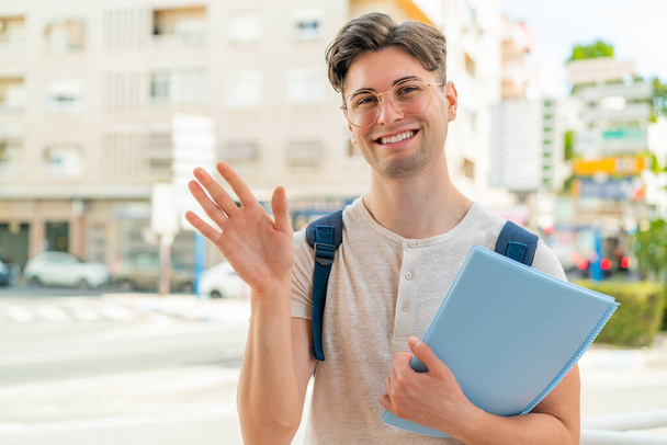 Young handsome student man at outdoors saluting with hand with happy expression - Photo, Image