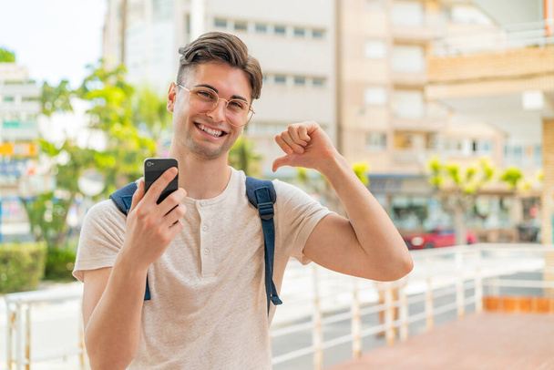 Jeune homme beau utilisant le téléphone mobile à l'extérieur fier et satisfait de soi - Photo, image