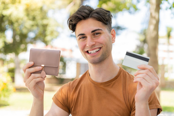 młody przystojny mężczyzna dzierżący portfel i kartę kredytową z szczęśliwy wyraz twarzy - Zdjęcie, obraz