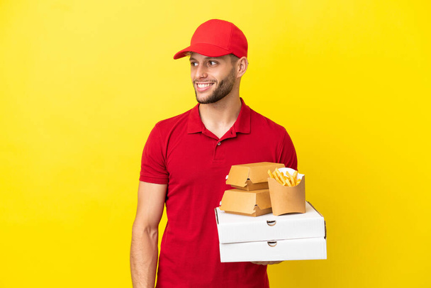 dostawca pizzy podnosząc pudełka po pizzy i hamburgery na odosobnionym tle patrząc na bok i uśmiechając - Zdjęcie, obraz