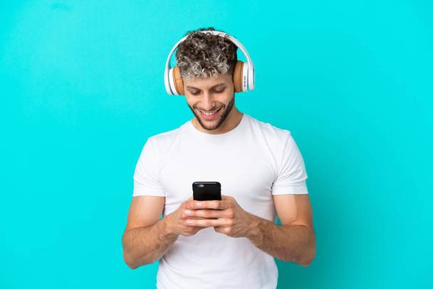 Young handsome caucasian man isolated on blue background listening music and looking to mobile - Photo, Image
