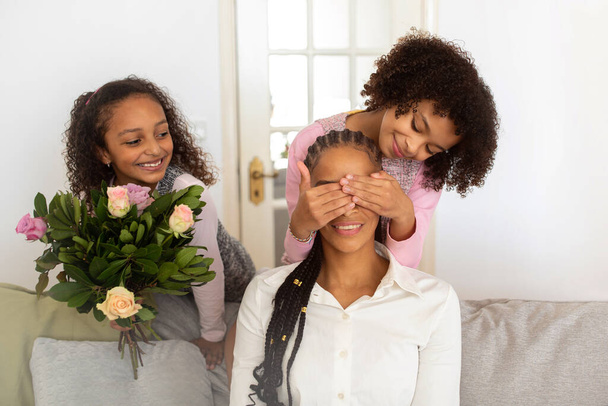 Prázdninová překvapení. Veselé černé dcery zavírající maminky oči a prezentující čerstvé květiny na ni doma, pózování v útulné domácí interiér. Šťastná rodina slaví Den matek nebo narozeniny - Fotografie, Obrázek