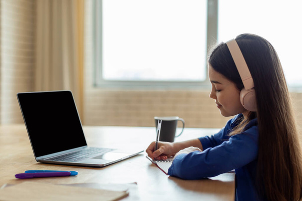 E-Learning-Angebot. Seitenansicht eines Schulmädchens, das mit leerem Bildschirm Notizen am Laptop macht, Kopfhörer trägt, online lernt und zu Hause am Schreibtisch Hausaufgaben macht. Attrappe - Foto, Bild
