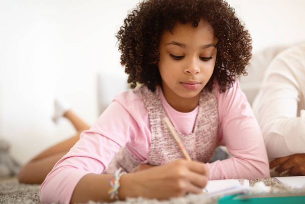 African American Preadolescent Girl Focused On Drawing With Watercolor Paint And Brush, Spending Time With Her Family Lying In Modern Living Room At Home. Weekend Leisure And Kid Hobby - Photo, Image