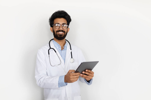 Handsome bearded friendly middle eastern young man in medical workwear doctor holding digital tablet, white wall background, copy space. Telemedicine, online appointment with doc, e-health - Zdjęcie, obraz