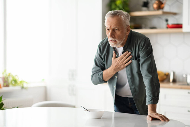 Een hartaanval. Oudere man die thuis pijn op de borst heeft, oudere man die een hartziekte heeft, zich niet lekker voelt, aan tafel staat in het keukeninterieur en borstkas wrijft, kopieerruimte - Foto, afbeelding