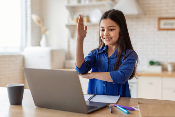 E-Learning Excellence. Glückliches Schulmädchen, das während eines Videotelefonats am Laptop die Hand hebt und zu Hause im Fernstudium Online-Unterricht hat. Moderne Bildung, digitales Klassenzimmer-Konzept - Foto, Bild