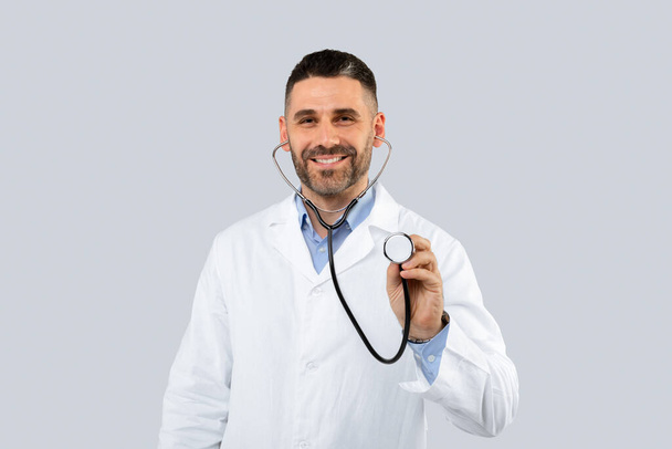 Friendly middle aged man doctor wearing white medical coat posing on light studio background, using stethoscope and smiling, copy space. Jobs and occupations - Photo, Image