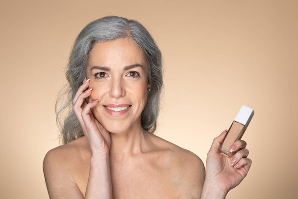 Πορτρέτο της όμορφης ηλικιωμένης Ευρωπαίας με καθαρό λαμπερό δέρμα, κρατώντας μπουκάλι μακιγιάζ ίδρυμα, θέτοντας σε μπεζ φόντο στούντιο. Σύσταση προϊόντος μακιγιάζ - Φωτογραφία, εικόνα