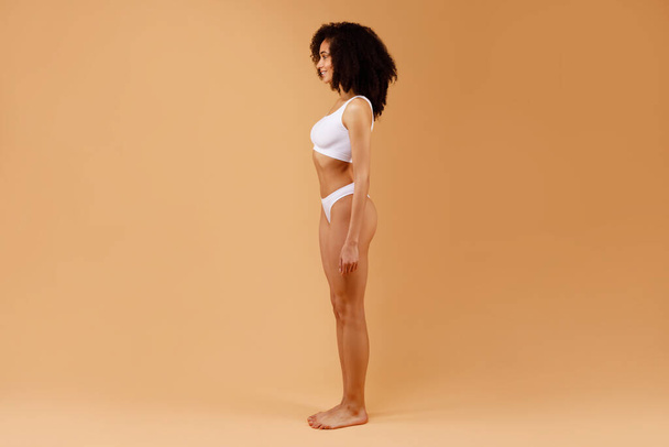 Zijaanzicht van slanke dame in wit ondergoed poseren geïsoleerd op beige studio achtergrond, sexy vrouw met fit slanke sportieve lichaam, volledige lengte, vrije ruimte - Foto, afbeelding