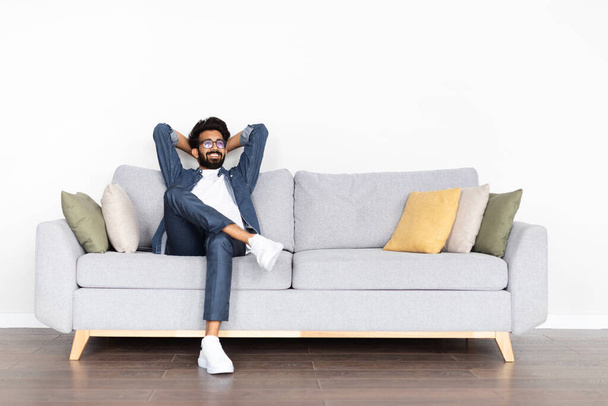Relaxed happy young eastern guy wearing eyeglasses and casual comfy outfit chilling alone on couch at home, looking at copy space and smiling, enjoying his new house, resting at weekend - Foto, afbeelding