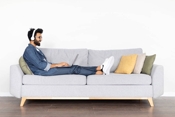 Cheerful millennial arab man in casual outfit wearing eyeglasses sitting on sofa over white wall, developer typing on laptop keyboard and smiling, using headphones, working from home, copy space - Photo, Image