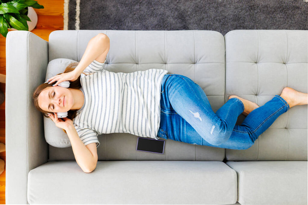 Peaceful woman in modern wireless headphones laying, relaxing on a comfortable couch, listening to music. Happy calm woman wearing headphones relaxing on a comfortable sofa, enjoy quality sound - Foto, afbeelding