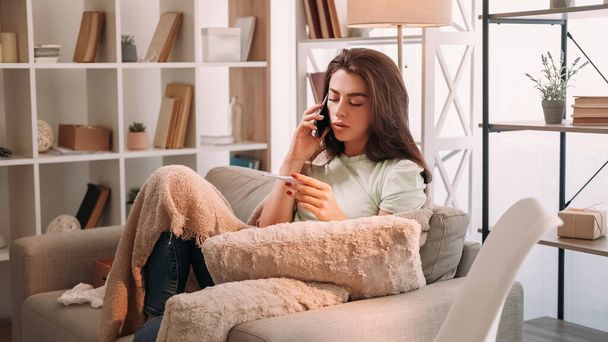 Virus disease. Sick woman. Online pharmacy. Tired lady covering plaid talking smartphone holding thermometer sitting sofa light room interior. - Photo, image