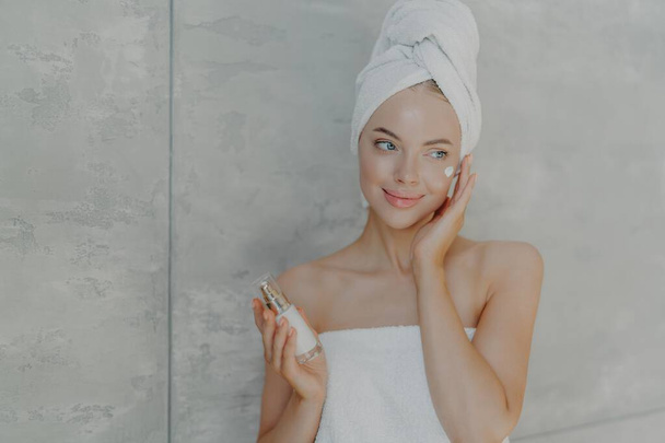Ικανοποιημένη γυναίκα εφαρμόζει λοσιόν ομορφιάς, πετσέτα στο κεφάλι, γυμνούς ώμους. Καλλυντικά, σπα έννοια σε γκρι φόντο. - Φωτογραφία, εικόνα
