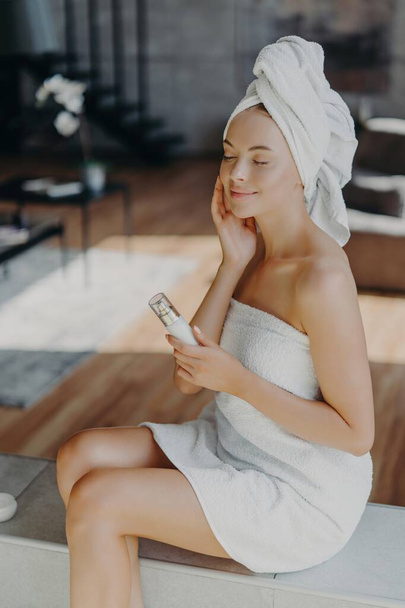Relaxed woman applies facial cream, wrapped in towel, in cozy home setting. Skincare and self-care concept. - Foto, Imagem