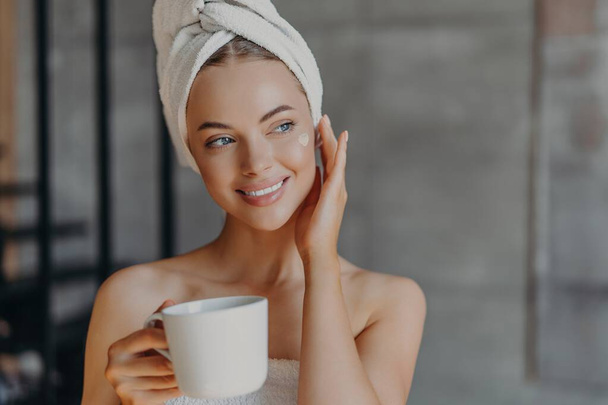 Pretty woman applies face cream, minimal makeup, wrapped in bath towel, enjoys coffee at home. Beauty, relaxation concept. - Photo, image