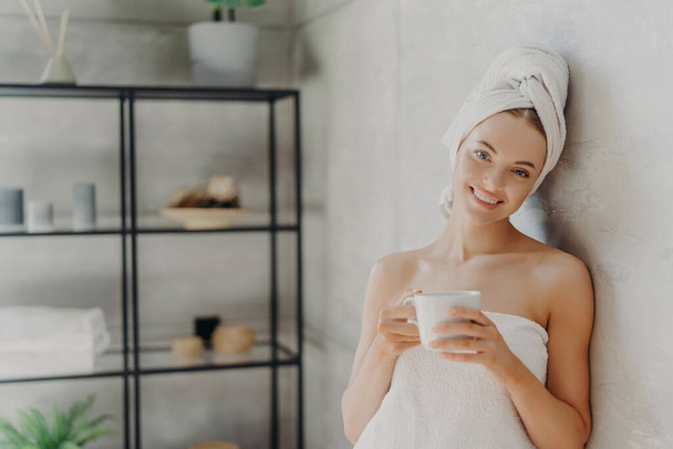 Mujer sonriente envuelta en toalla, bebe café, disfruta del spa interior. Bienestar, concepto de relajación. - Foto, Imagen