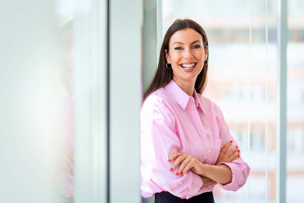 Zdjęcie portretowe atrakcyjnej bizneswoman patrzącej w kamerę i uśmiechającej się stojącej z rękami skrzyżowanej w nowoczesnym biurze. - Zdjęcie, obraz
