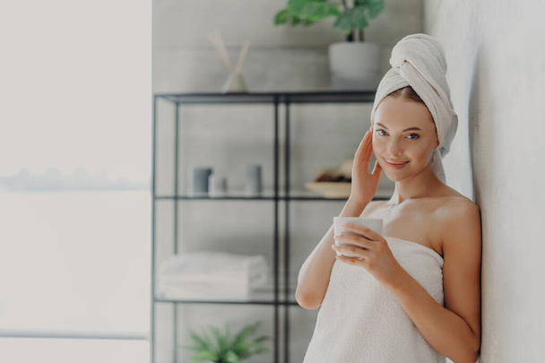Pleased woman enjoys spa with glowing skin, towel-wrapped, drinks tea in cozy bathroom. Beauty and relaxation. - Photo, Image