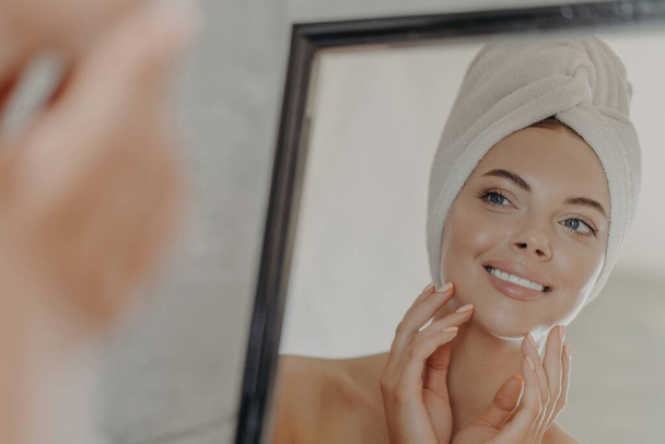 Adorable woman admires reflection, touches healthy skin, towel on head. Beauty routine. - Photo, Image