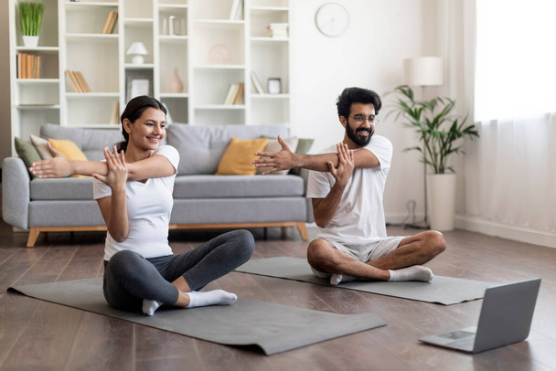 Fröhliches indisches Paar, das zu Hause Yoga praktiziert, lächelnde östliche Ehepartner, die auf Laptop-Bildschirm schauen und sich auf Fitnessmatte strecken, Online-Triining-Kurs genießen, gemeinsam im Wohnzimmer trainieren - Foto, Bild