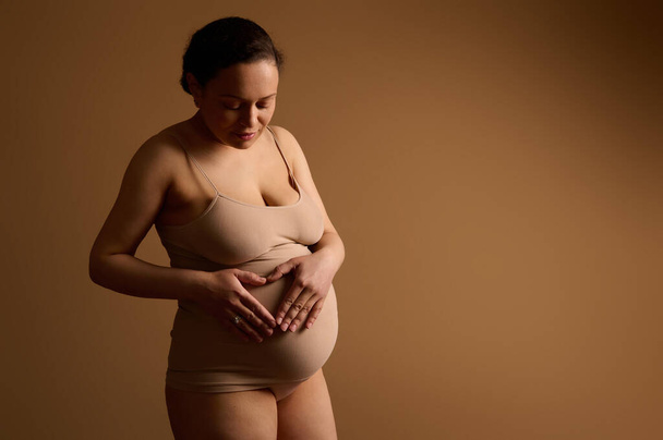 Multi etnische prachtige jonge gravid verwachting vrouw in ondergoed, het vormen van hartvormige handen op haar zwangere buik, uiten van gelukkige positieve emoties, genieten van haar zwangerschap, geïsoleerde beige achtergrond - Foto, afbeelding
