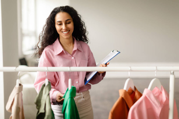 Fashion Retail Success. Confident Entrepreneur Woman Managing Clothing Collection Posing With Clipboard In Hand, Standing Near Clothes Rack In Trendy Store Indoors. Small Business, Entrepreneurship - Photo, Image