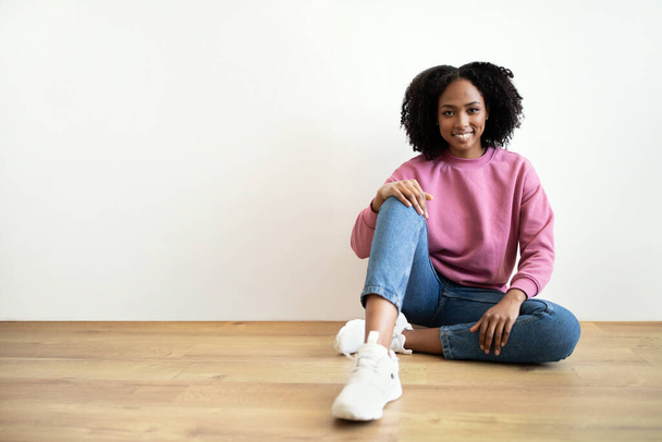 Happy millennial african american curly lady in casual sitting on floor in living room interior, on white wall. Rest, relax, break and lifestyle at home alone, ad and offer - Фото, изображение